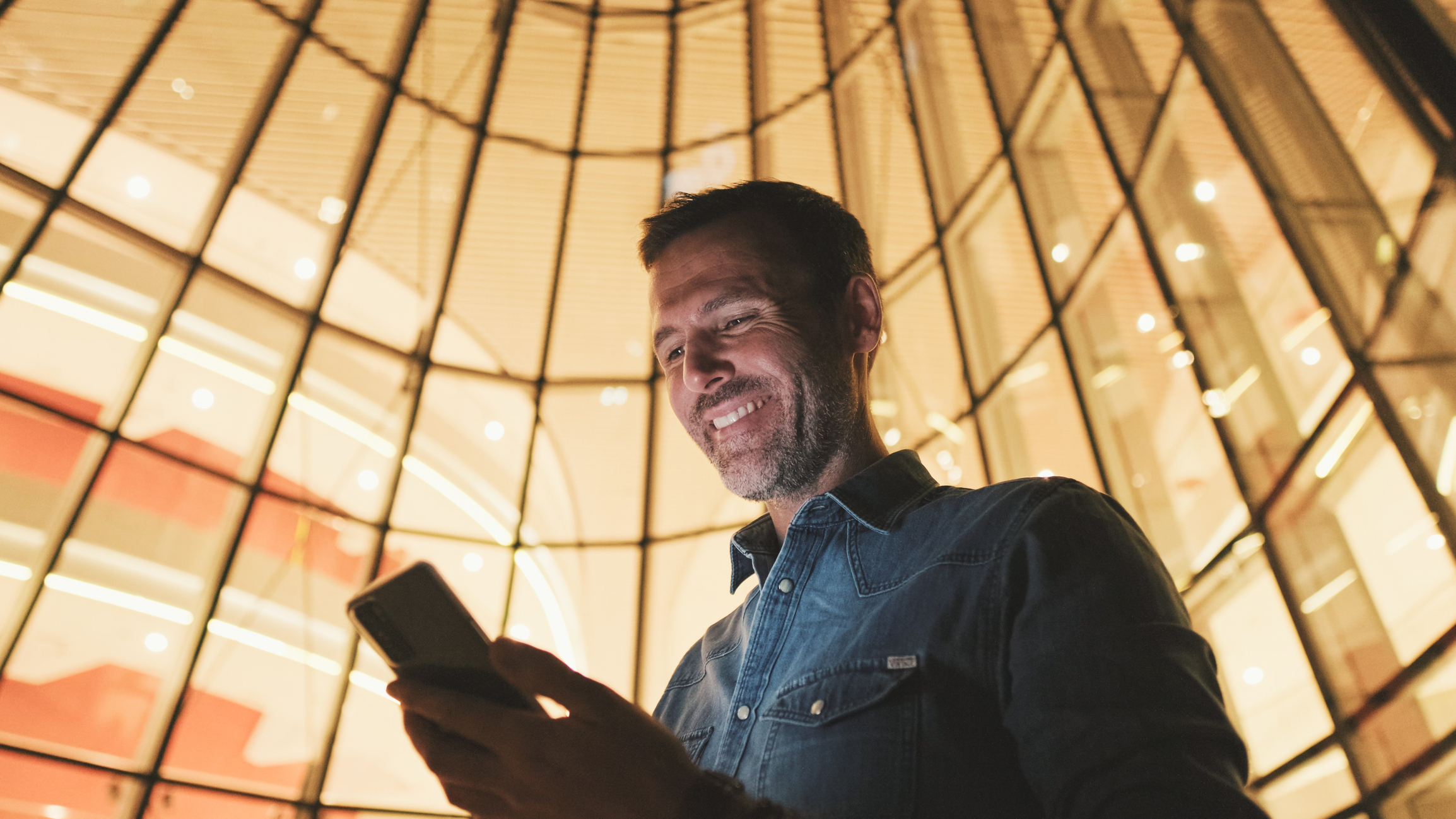 Man smiling and looking down at phone, against a background of warm yellow windows.