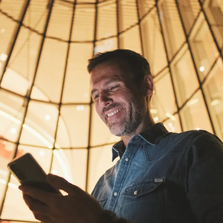 Man smiling and looking down at phone, against a background of warm yellow windows.