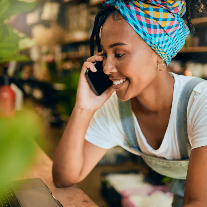 A woman wearing overalls and a brightly-coloured headscarf, using a phone and laptop.