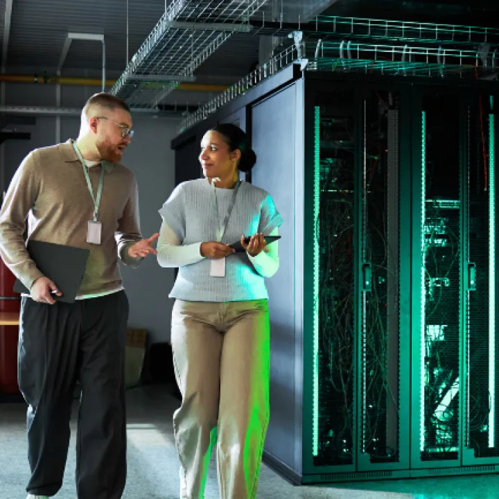 Two staff walk through a data centre server room, talking while holding a tablet and folder, with tall server racks and network cables visible on the right.