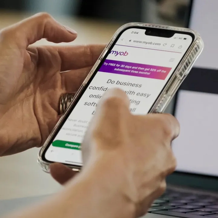 Close up image of hands holding a smart phone while using the MYOB website
