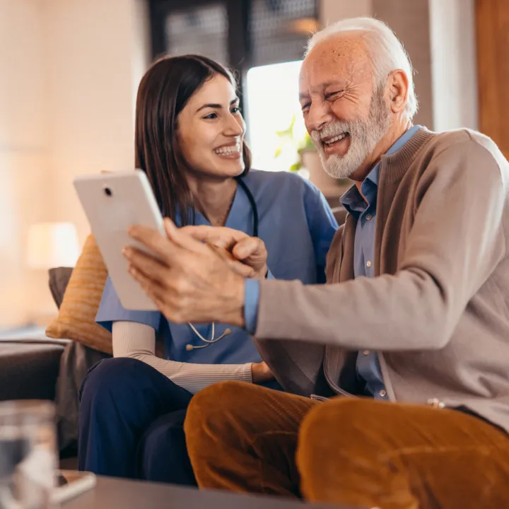 Nurse assisting senior man using digital tablet at home, both happily chatting.