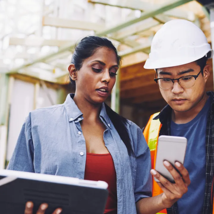 Construction industry manager sharing information by showing her colleague her smartphone. Her colleague is a man wearing a high-vis vest and hardhat.