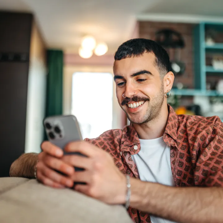 A young man with a moustache and nose rings sitting in his living room looking at information on a smartphone.